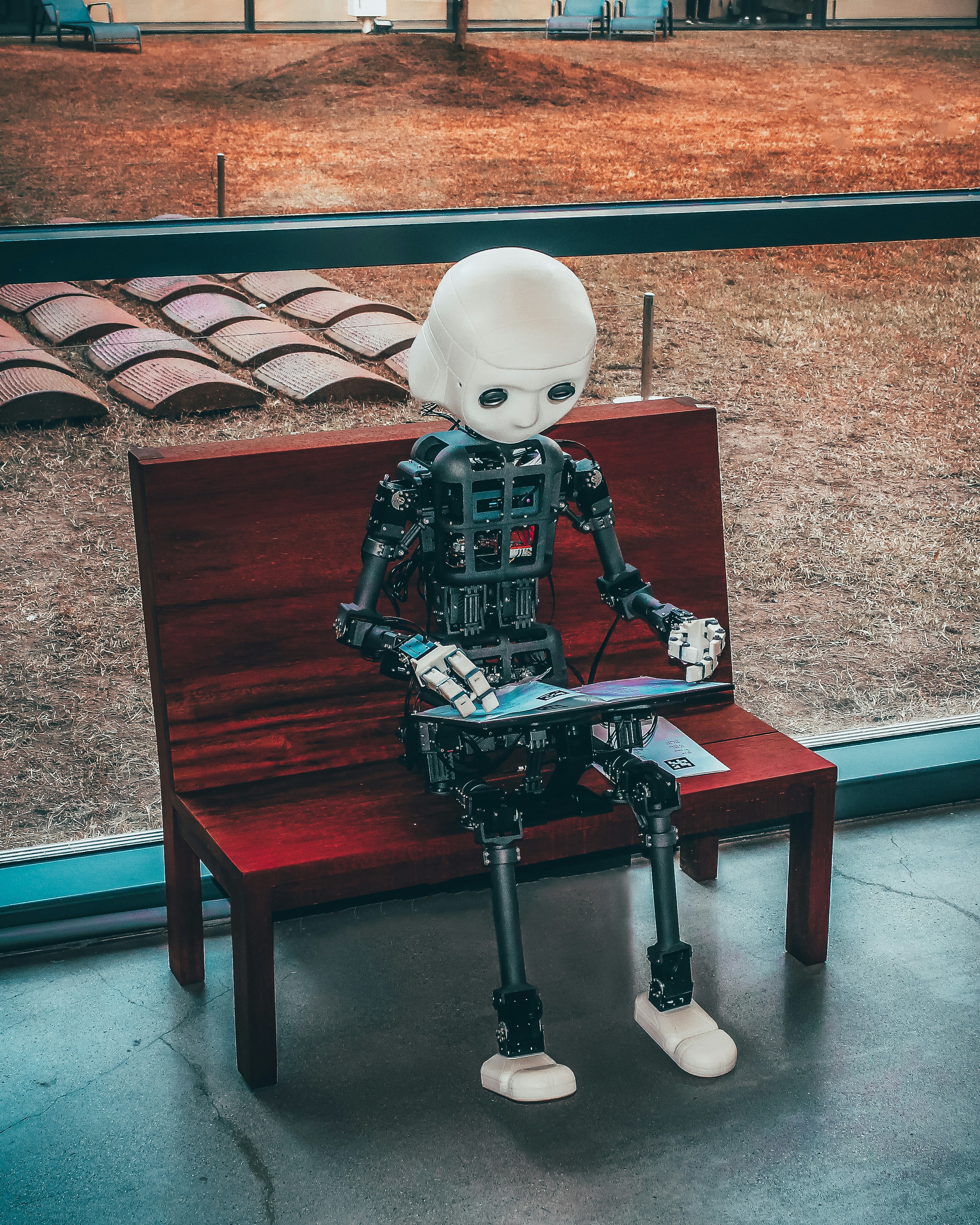 A humanoid robot seated on a bench, leaning forward and looking down at a screen in its hands.