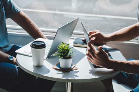 Two people at a round table with laptops, coffee, and a plant, discussing work in a bright office.