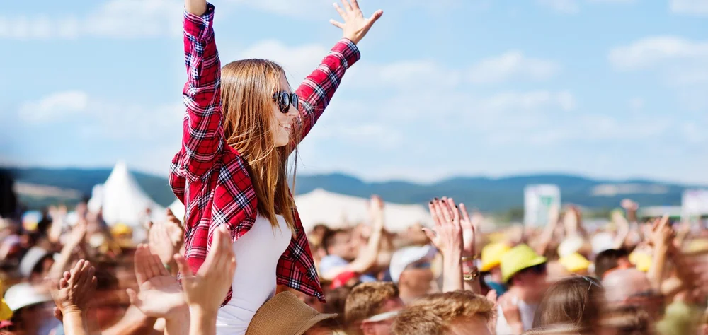 Girl sitting on someone’s shoulders at a festival with arms raised, enjoying the crowd