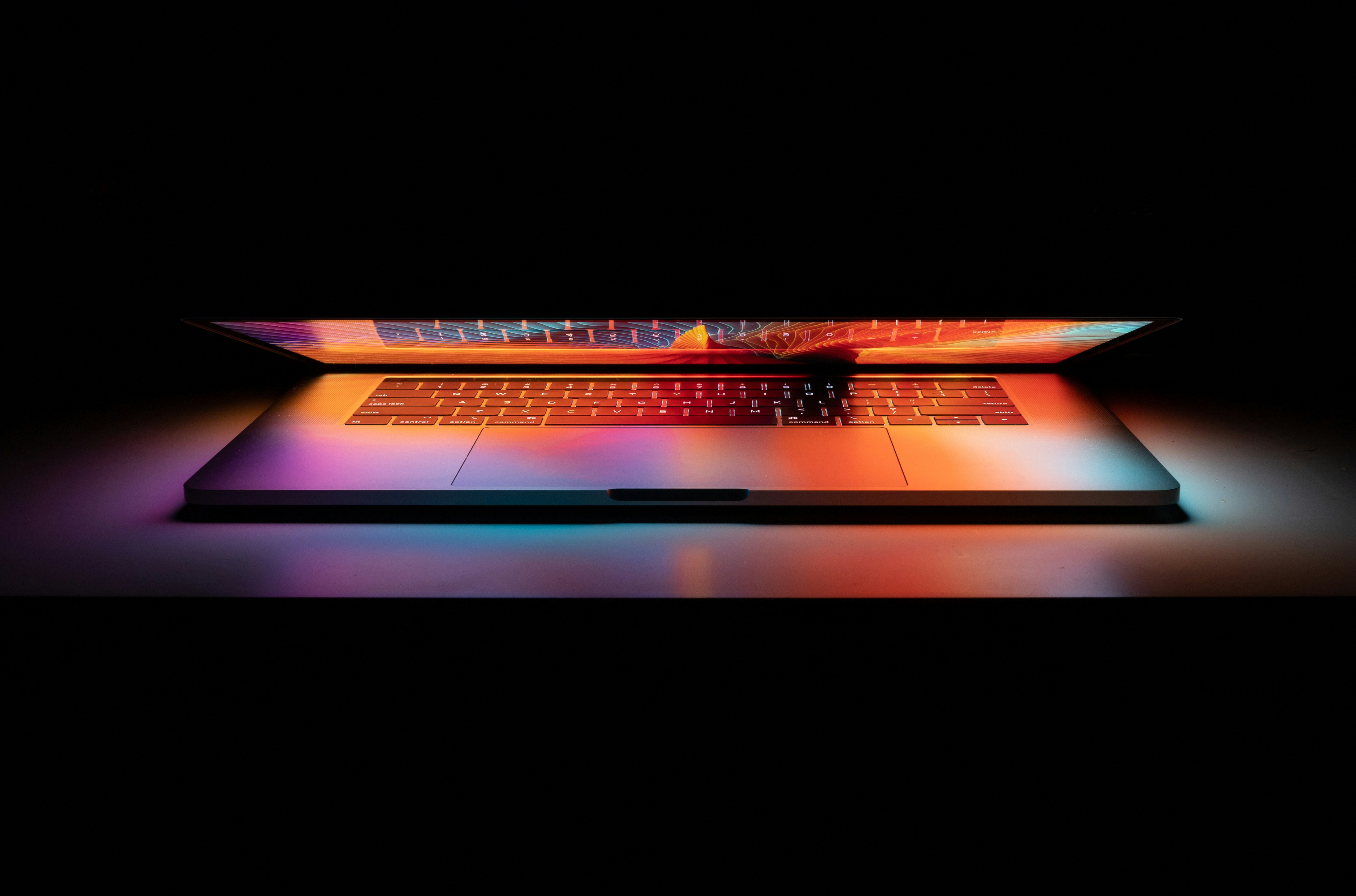 A partially open laptop illuminated by vibrant orange, pink, and blue light against a dark background.